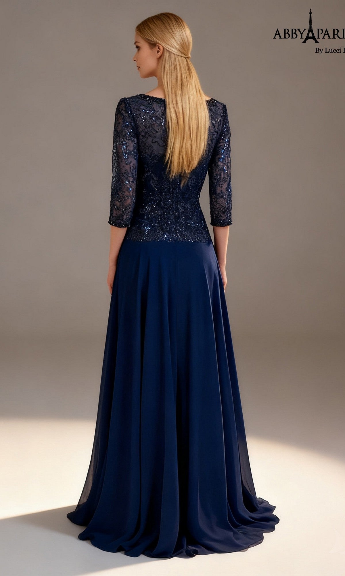A woman stands facing away, wearing the Abby Paris 96099 long formal dress in navy blue. The lace bodice features three-quarter sleeves and is adorned with sequin-embellished detailing for a touch of sparkle, while the long, flowing chiffon A-line skirt completes the elegant look. Her straight blonde hair falls loosely down her back.